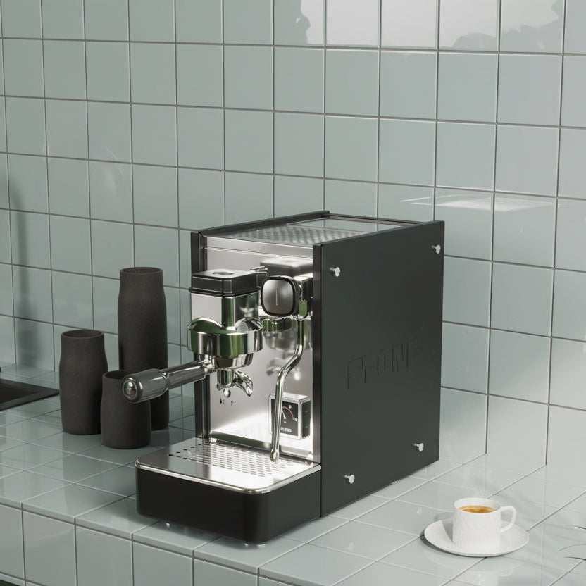 A modern espresso machine with a matte black body and polished stainless steel front, set on a light gray tiled kitchen counter next to a cup of coffee.